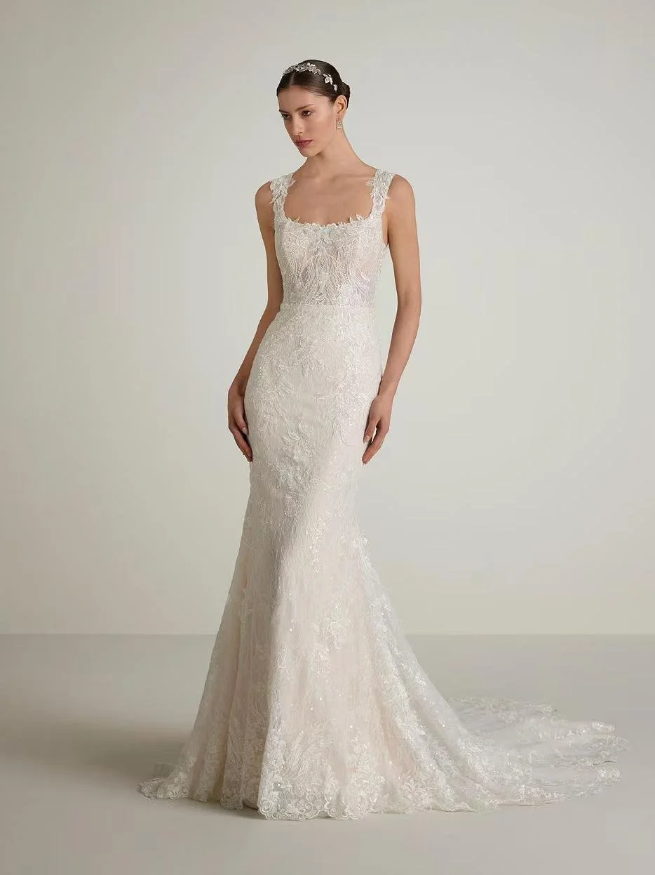 A woman wearing a white lace wedding dress with a fitted bodice and a train, standing against a plain background.
