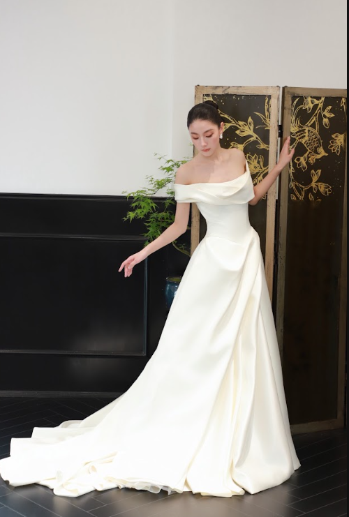 A woman in an off-the-shoulder white wedding gown standing indoors near a decorative folding screen.
