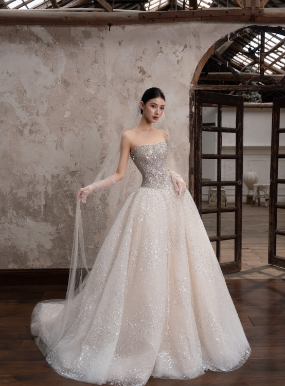 A woman in an elegant, sparkly strapless wedding gown with a full skirt, standing inside a rustic building.