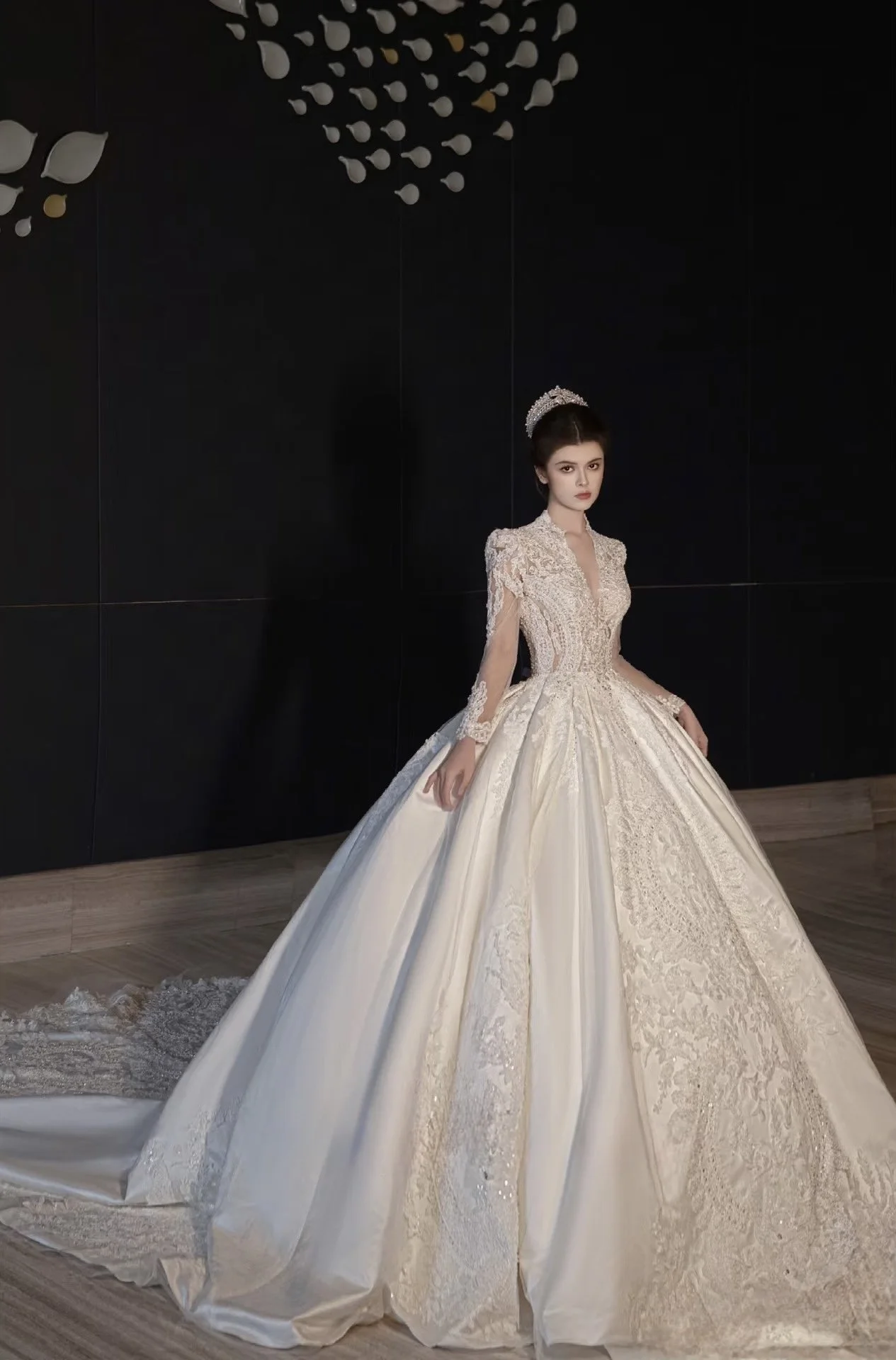 A woman wearing a detailed, elegant white wedding gown with lace and embroidery, standing in front of a black wall with decorative elements.