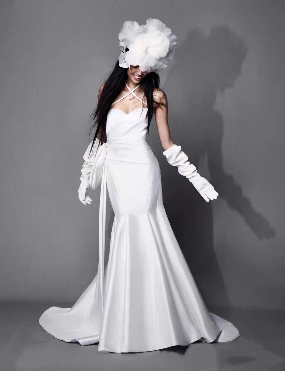 Woman in a long white gown with gloves, smiling, and a headpiece made of large white fabric flowers, standing against a gray background.