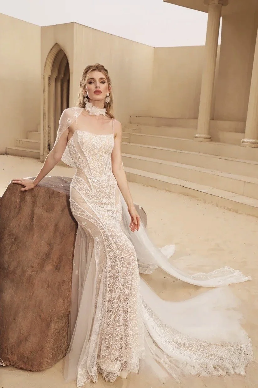 A woman in a white lace wedding dress with a train, standing next to a large brown rock in a neutral-toned, minimalist setting with a staircase and an arched doorway in the background.
