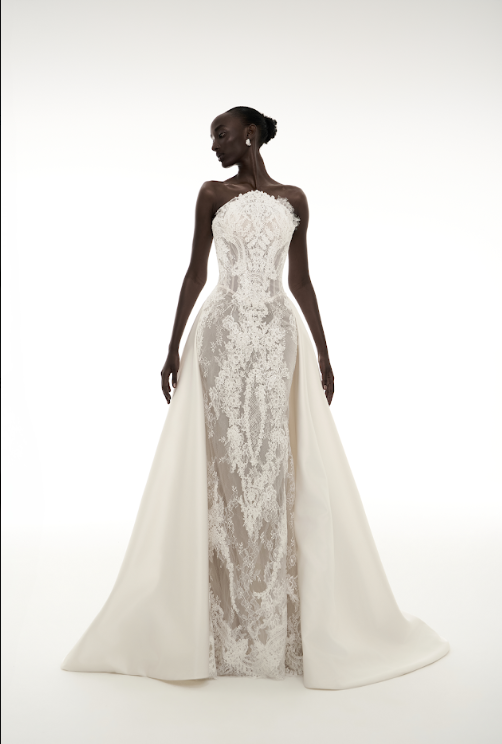 A woman in a white wedding dress with lace details and a high slit, standing against a plain white background.
