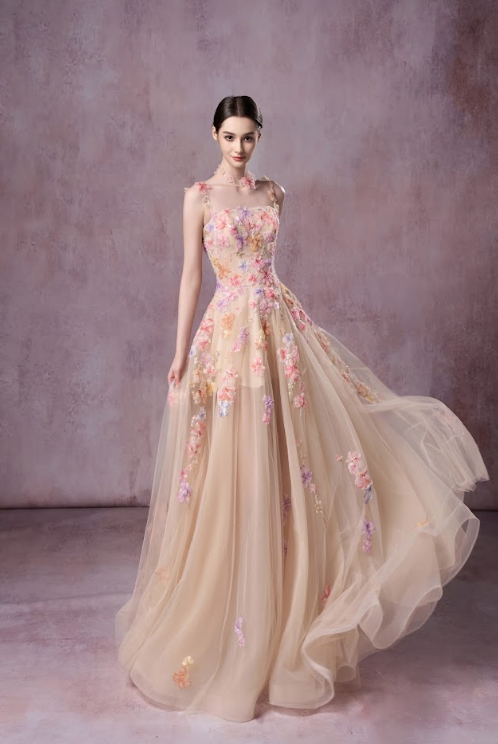 A woman in a long, pink floral embroidered gown with a full tulle skirt, standing against a muted pink textured background.