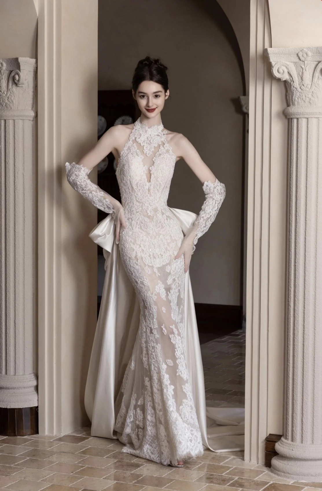 A woman in an elegant white lace wedding gown with long lace gloves, standing in a doorway with classic columns.