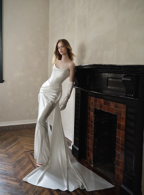 A young woman with long wavy hair sitting on a black bench against a plain beige wall, wearing a strapless satin white dress and matching gloves.