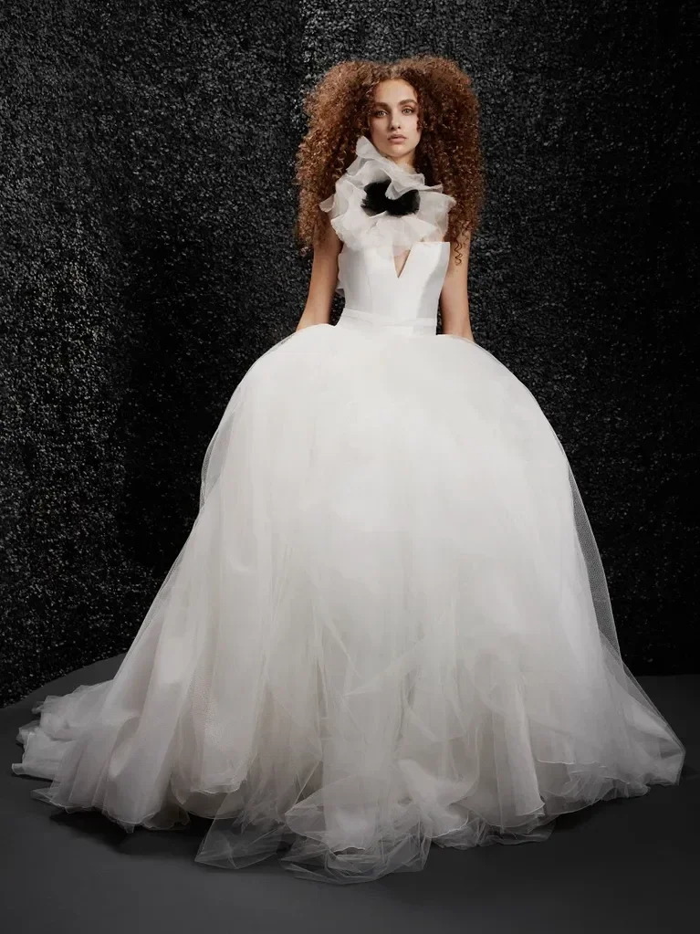 A woman wearing a white bridal gown with a voluminous skirt and large ruffled collar, standing against a dark, textured background.
