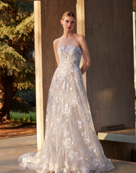 A woman in a strapless white wedding gown with lace detailing, standing outdoors near a concrete structure, with trees and sunlight in the background.