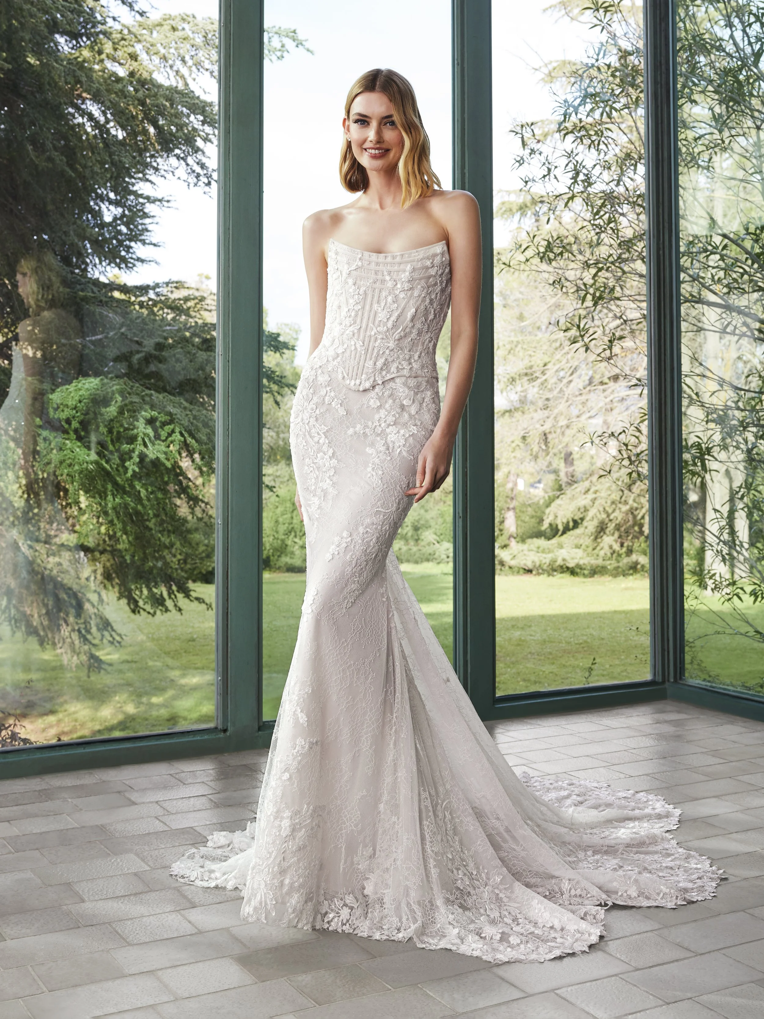 A woman in a white lace wedding gown standing indoors near large windows with a garden view outside.