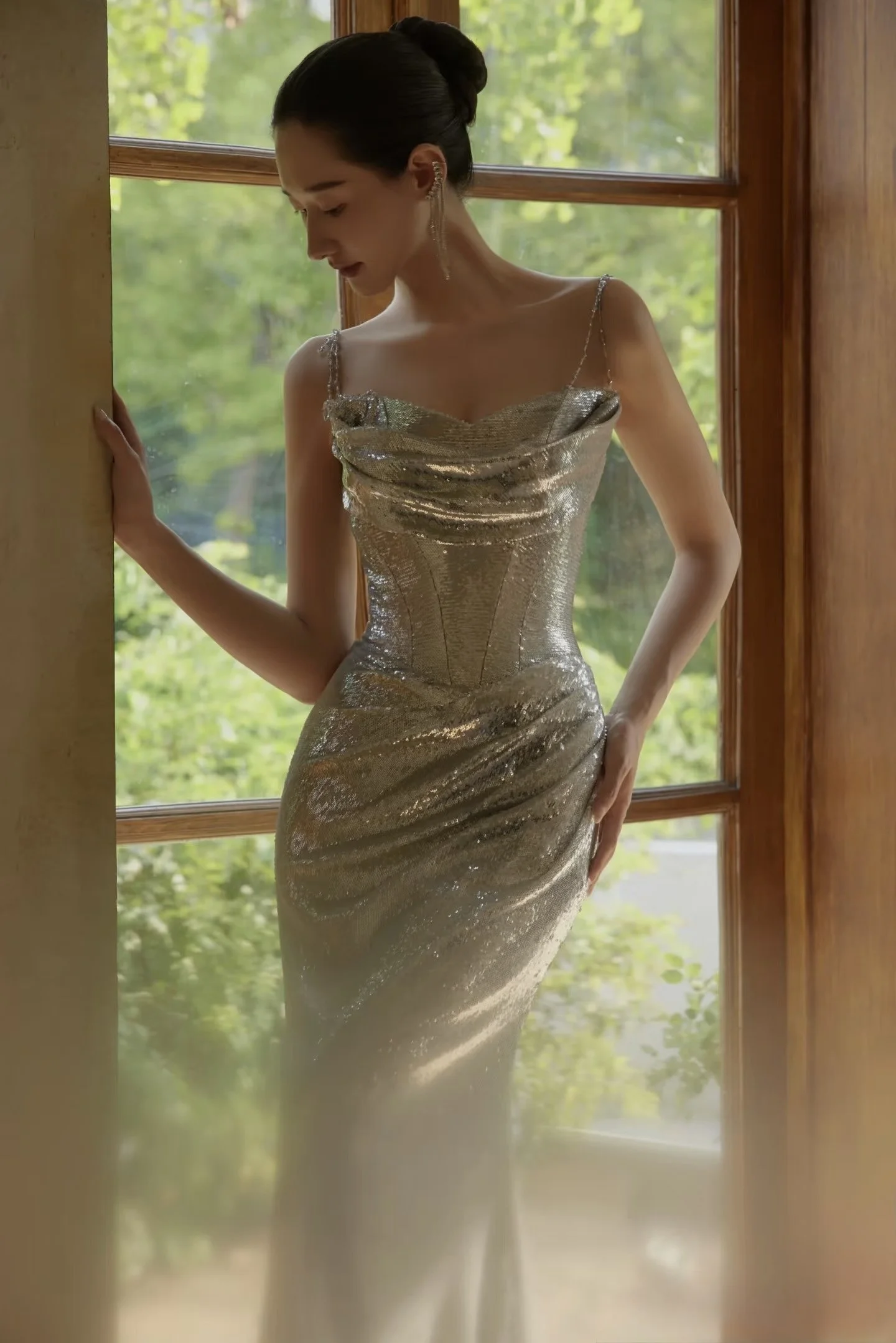 A woman in a shiny, gold, floor-length gown with thin straps, standing by a wooden-framed glass door, with her head turned downward and hair styled in an elegant updo, in a bright room with green trees visible outside.