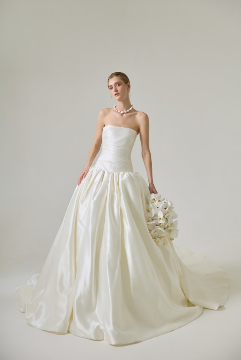 A woman in a strapless white wedding gown holding a bouquet of white flowers, standing against a plain light-colored background.