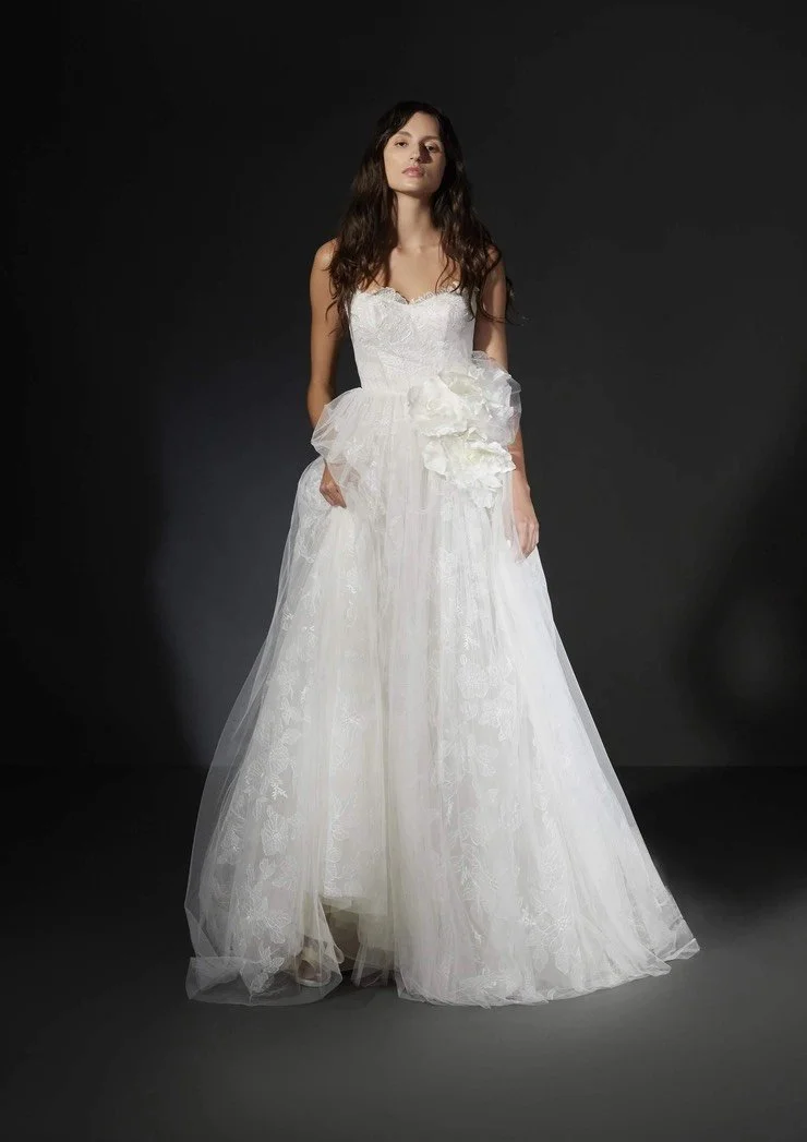 Woman wearing a white wedding dress with lace details and floral accents, standing against a dark background.