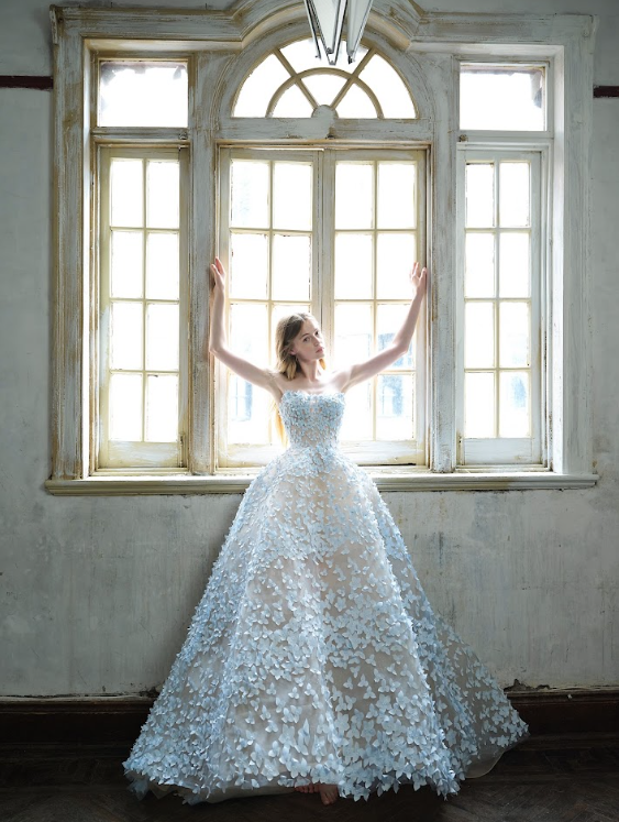 A woman wearing a strapless white wedding gown with floral appliqué details stands in front of large, old-fashioned windows with her arms raised.