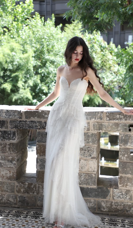 Young woman in a strapless white wedding dress standing outdoors on a stone balcony, looking down with a serious expression.