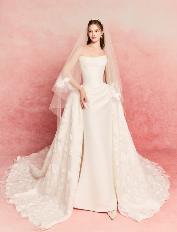 A woman in a white wedding dress with a long train and veil, standing against a pink background.