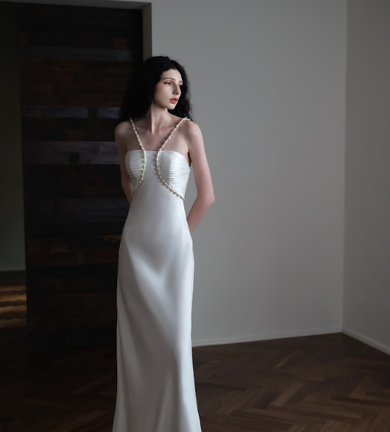 A woman in a white, sleeveless dress with thin straps stands in a room with dark wood flooring and white walls, looking to her left.