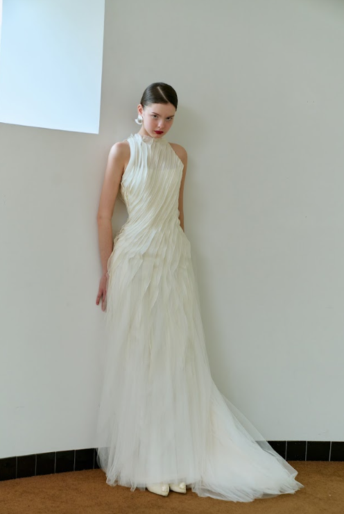 A woman wearing a long, elegant, ivory pleated gown standing against a plain light-colored wall.