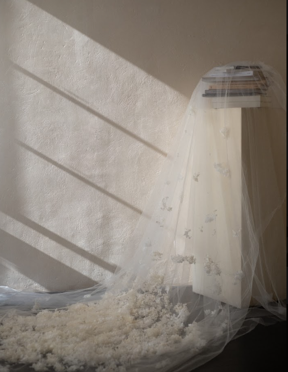 A wooden nightstand covered with a semi-transparent fabric, possibly a sheer curtain, with white fluffy material on the floor and a plain wall in the background, illuminated by natural sunlight and casting shadows.