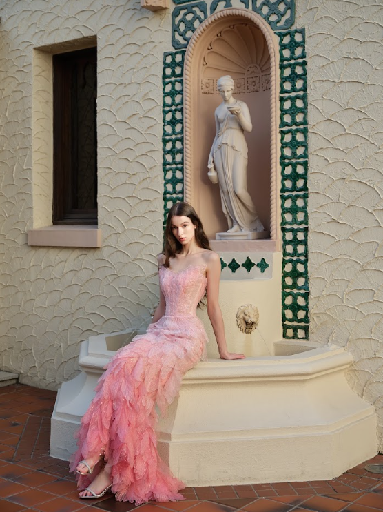 A young woman in a pink, feathered gown sitting on a curved architectural feature in front of a decorative wall with a niche containing a classical statue. The wall has textured cream-colored surface and a small window to the left.