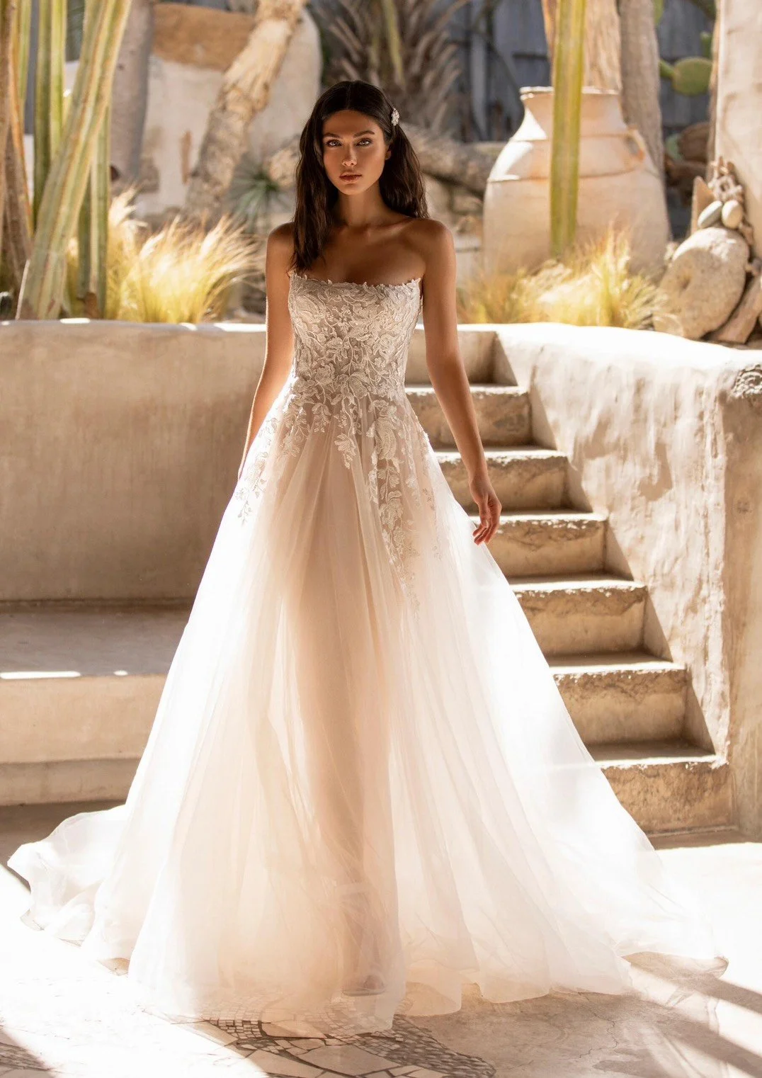 A woman wearing a strapless wedding dress with lace bodice and flowing tulle skirt standing outdoors on stone steps with desert plants and pottery in the background.