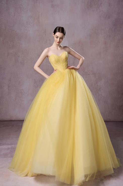 A woman in a strapless, yellow ball gown with a full, layered skirt, standing against a plain, muted background.