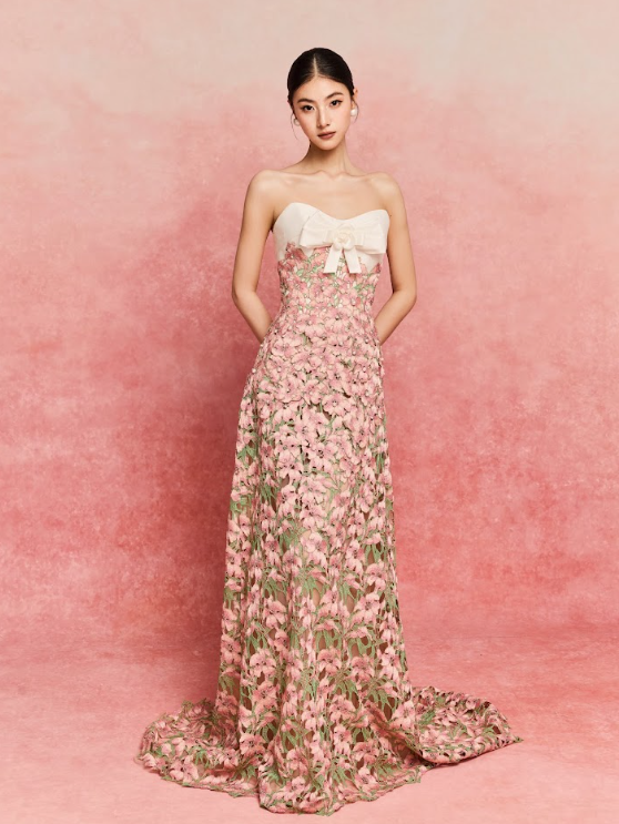 A young woman in a strapless pink floral gown with a white bow on the top, standing against a pink textured background.