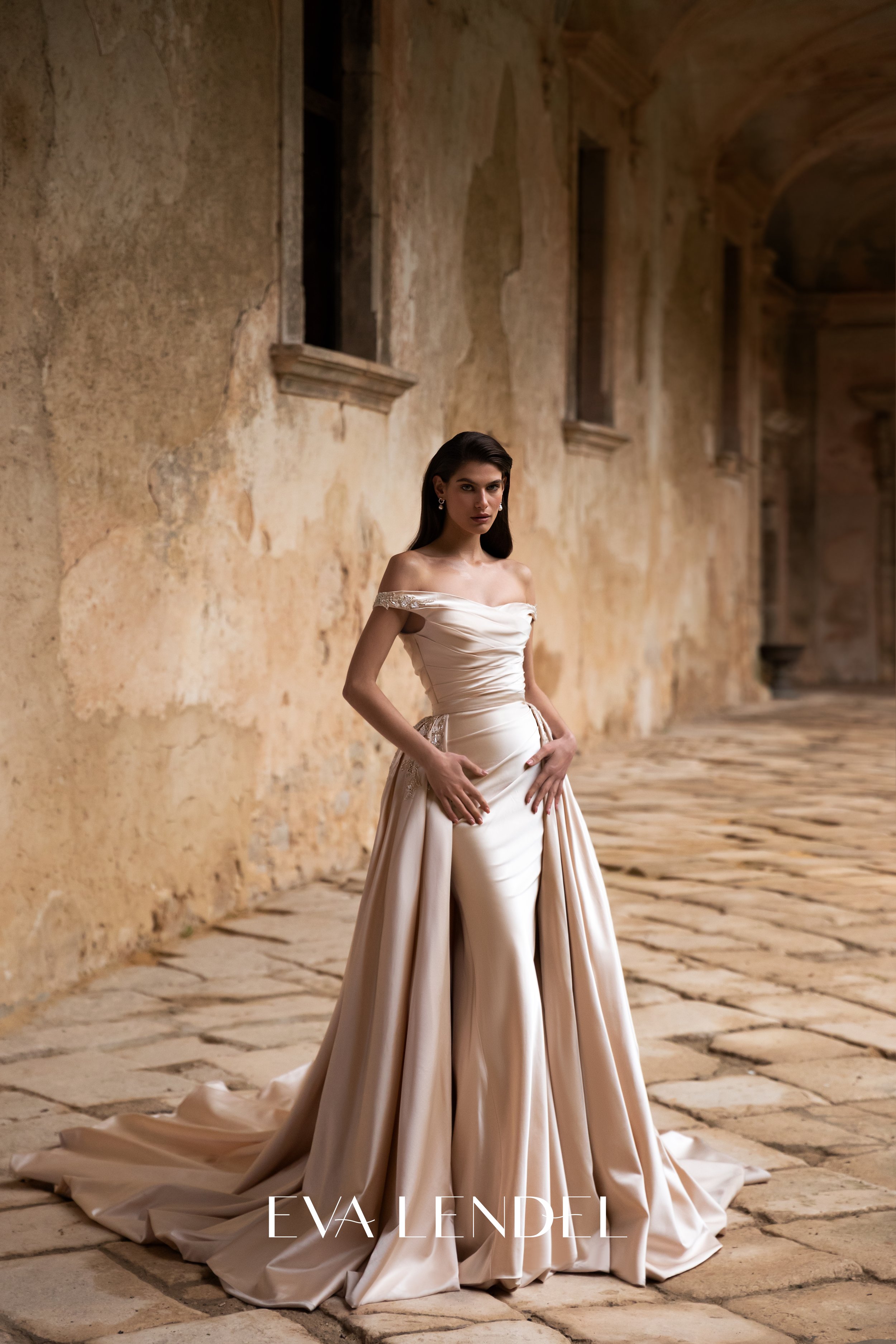 A woman wearing a luxurious off-the-shoulder satin gown with a full skirt, standing in an old, weathered stone corridor with archways and open windows. The image features text that says 'EVA LENDEL' at the bottom.