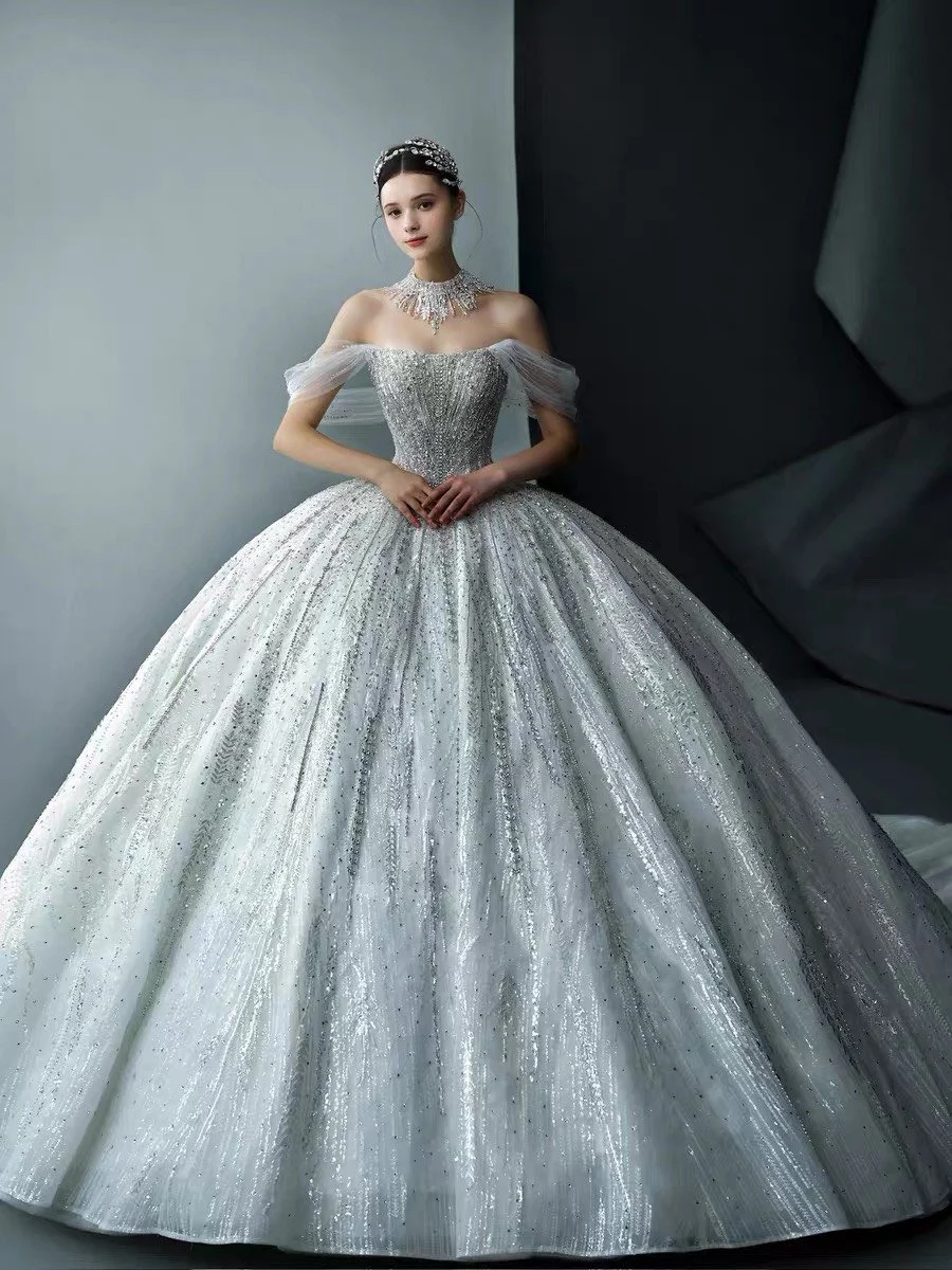 A young woman in an elegant silver, off-the-shoulder, ball gown with a full, glittery skirt, adorned with jewelry, standing against a gray and black background.