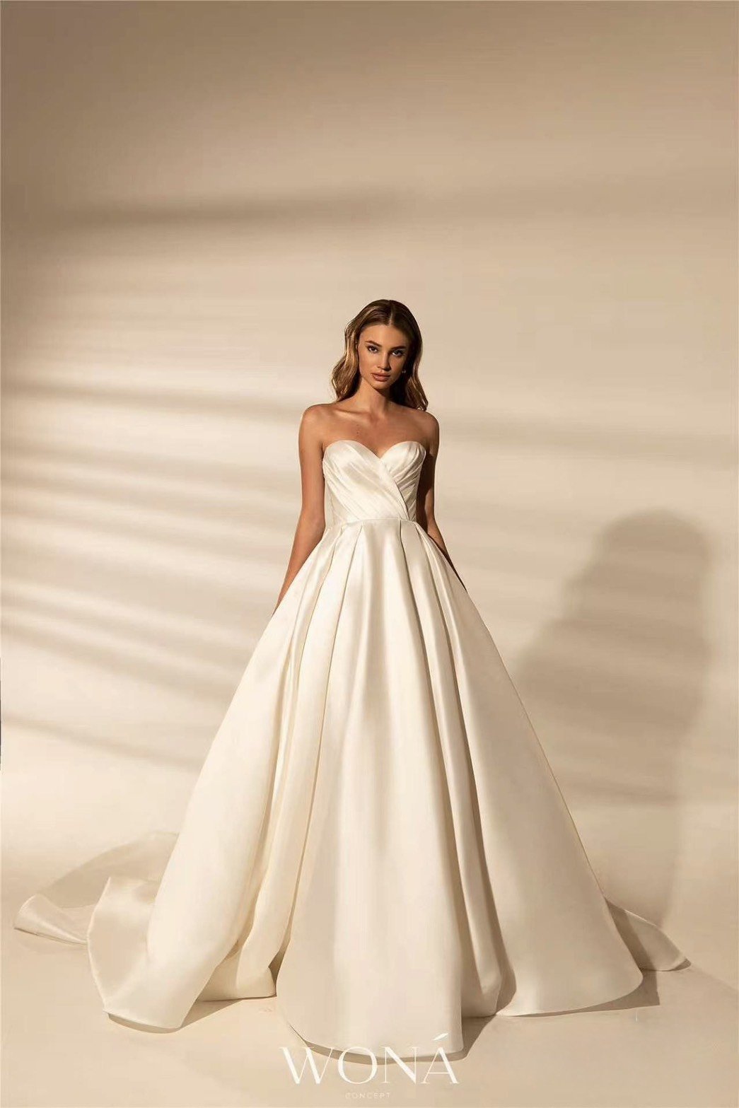 A woman wearing a strapless white wedding gown with a full skirt, standing against a neutral background with soft lighting.