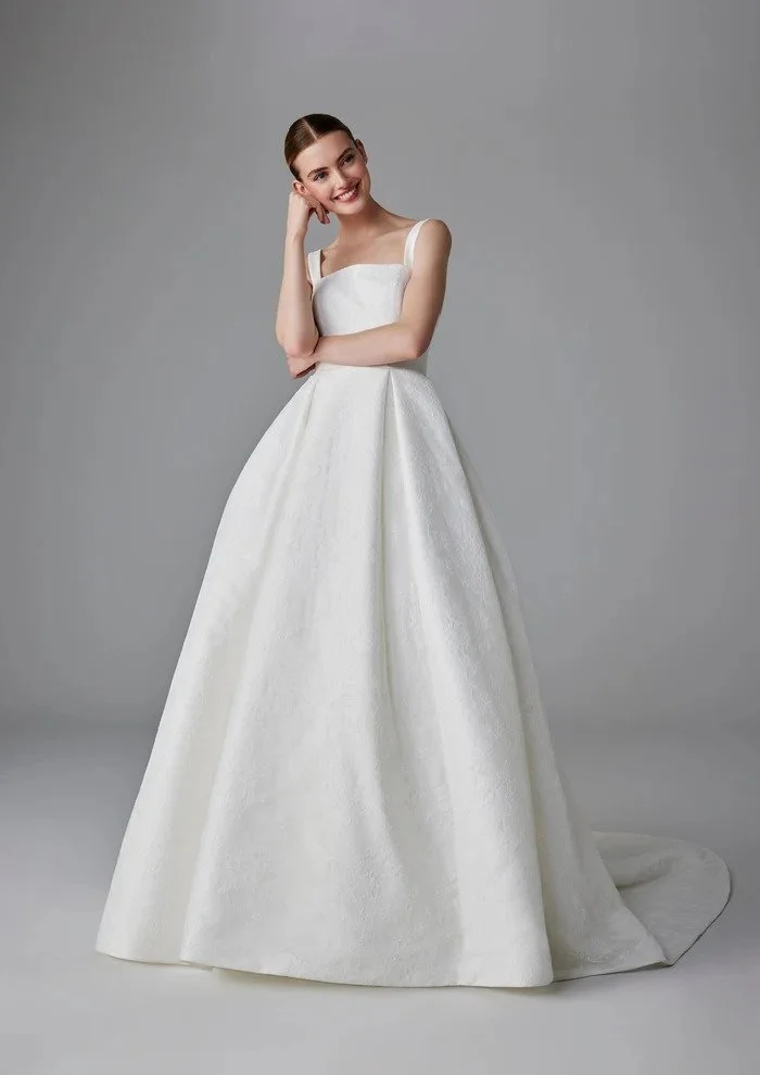 A woman in a white wedding dress standing against a plain gray background, smiling and touching her hair.