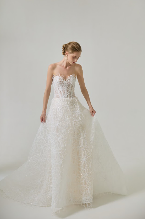 A woman in a strapless white wedding dress with lace detailing stands against a plain gray background, looking down and holding the sides of her dress.