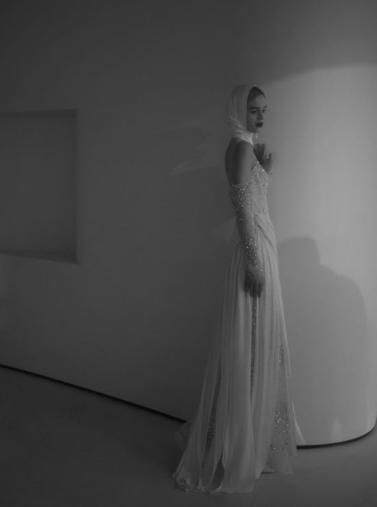A woman in a long, elegant dress with intricate beadwork, standing against a plain white wall in a minimalist setting, with her hand touching the wall and hair covered with a headscarf.