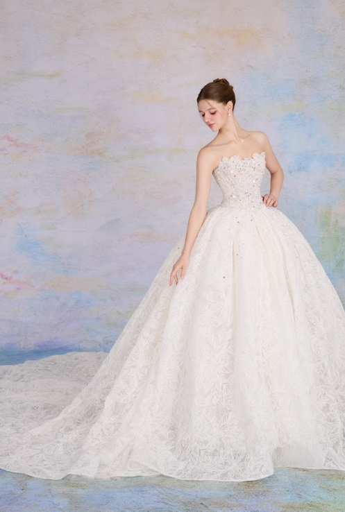 A young woman in a white wedding gown with lace details, standing against a pastel-colored background.