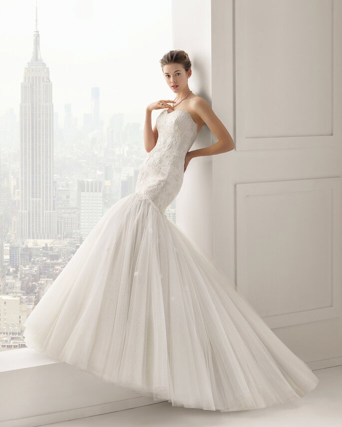 A woman in a white wedding gown standing in a bright room with a city skyline in the background, including the Empire State Building.
