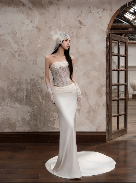 A woman in a wedding dress standing in a rustic interior with a textured wall and wooden floor, wearing a lace strapless gown, lace gloves, and a tulle veil.