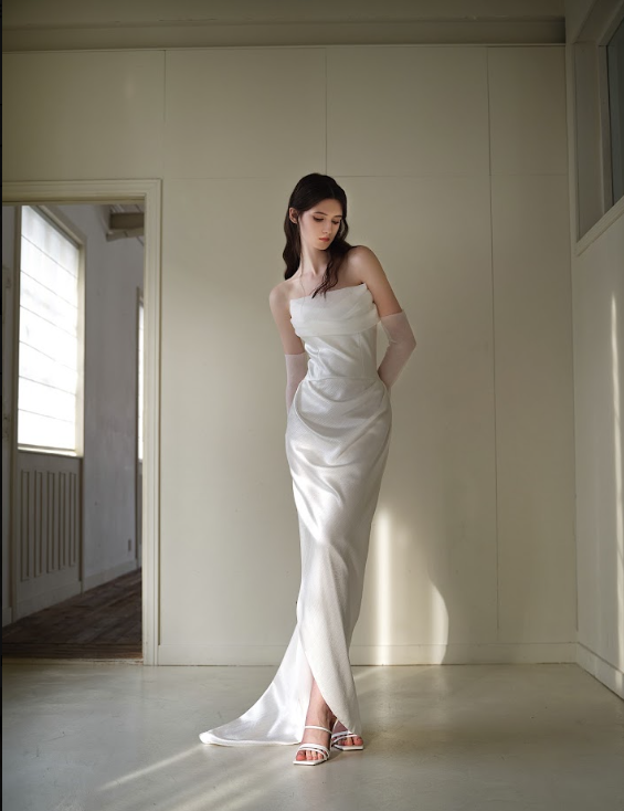 A woman in a white satin gown with off-the-shoulder sleeves standing in a minimally decorated room.
