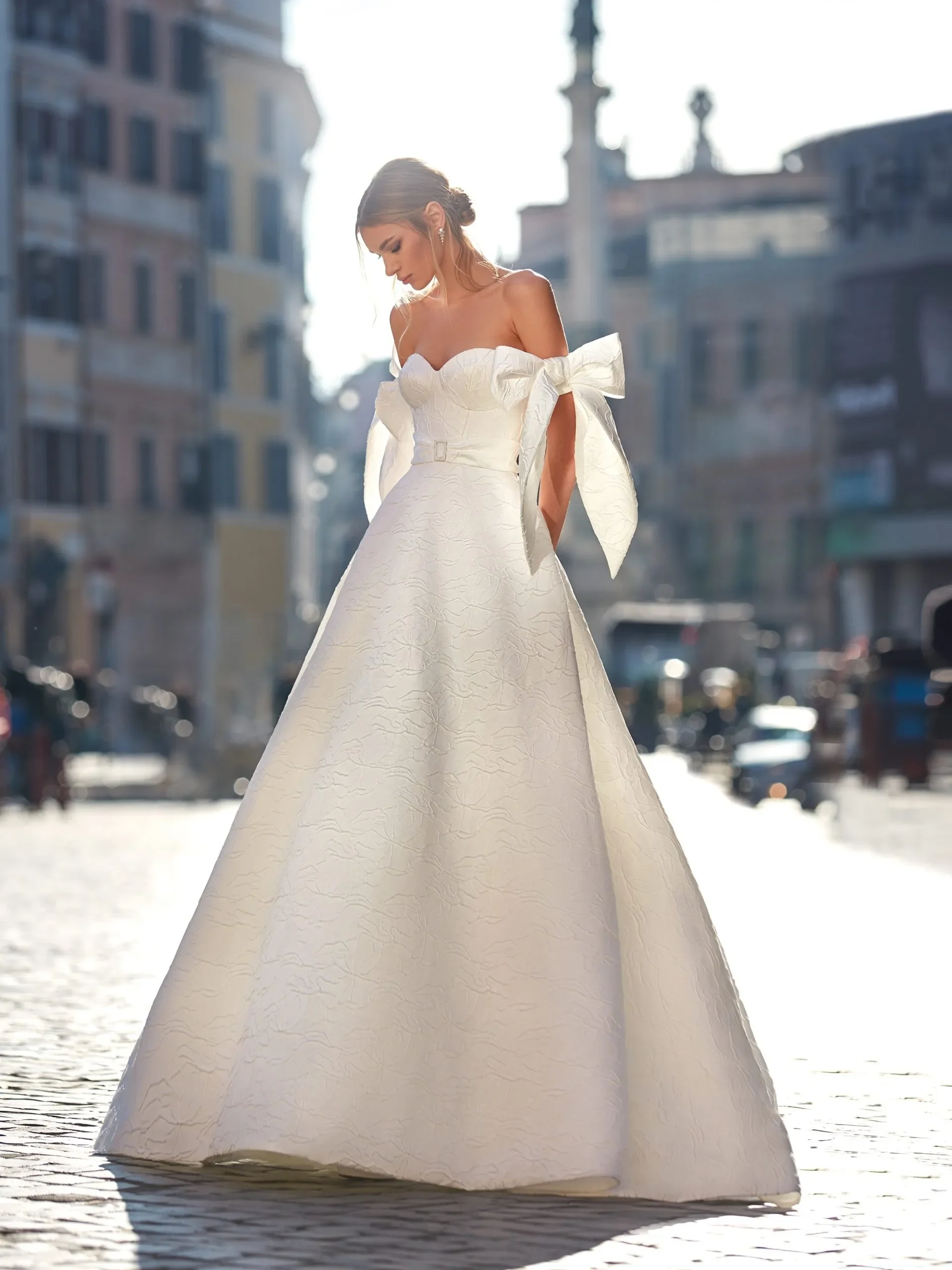 A woman in a white wedding dress standing outdoors on a cobblestone street, with blurred buildings in the background, illuminated by sunlight.