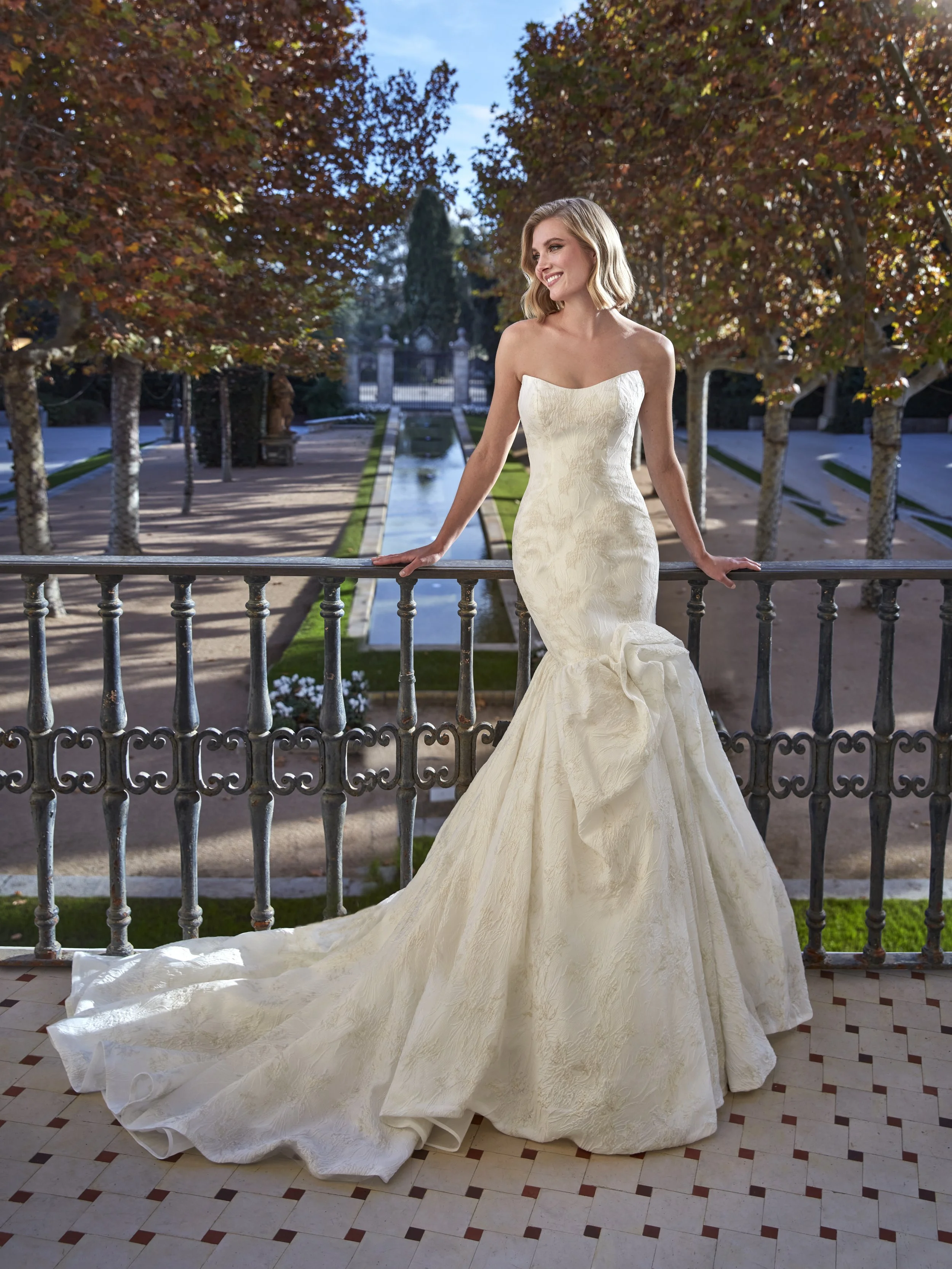 A woman in a strapless white wedding gown standing outdoors on a balcony with a metal railing, trees with orange and green leaves, a fountain, and a gated entrance in the background.