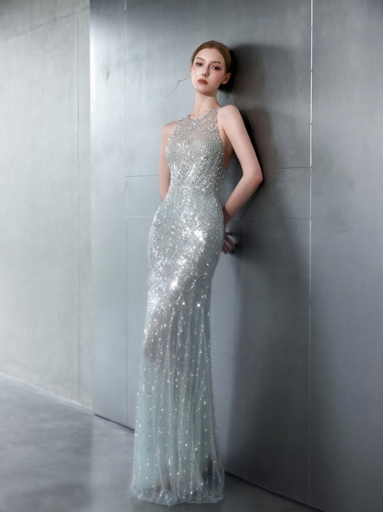 A young woman in a sparkly, silver, sleeveless evening gown standing against a gray metallic wall.