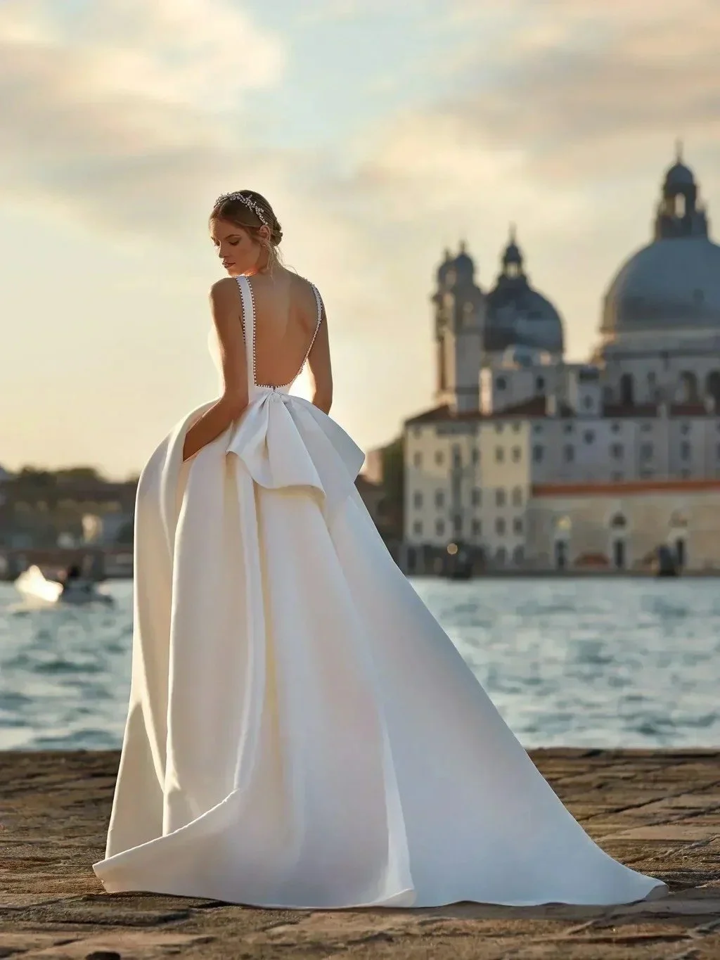A bride in a white wedding gown standing by water during sunset with a historic European city and domed buildings in the background.