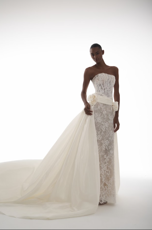 A woman in a strapless white lace wedding dress with a long train and floral sash, standing against a plain white background.