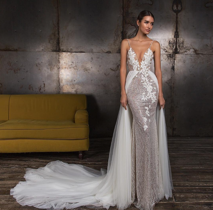 A woman in a detailed wedding dress standing indoors with a yellow couch and metal wall behind her.