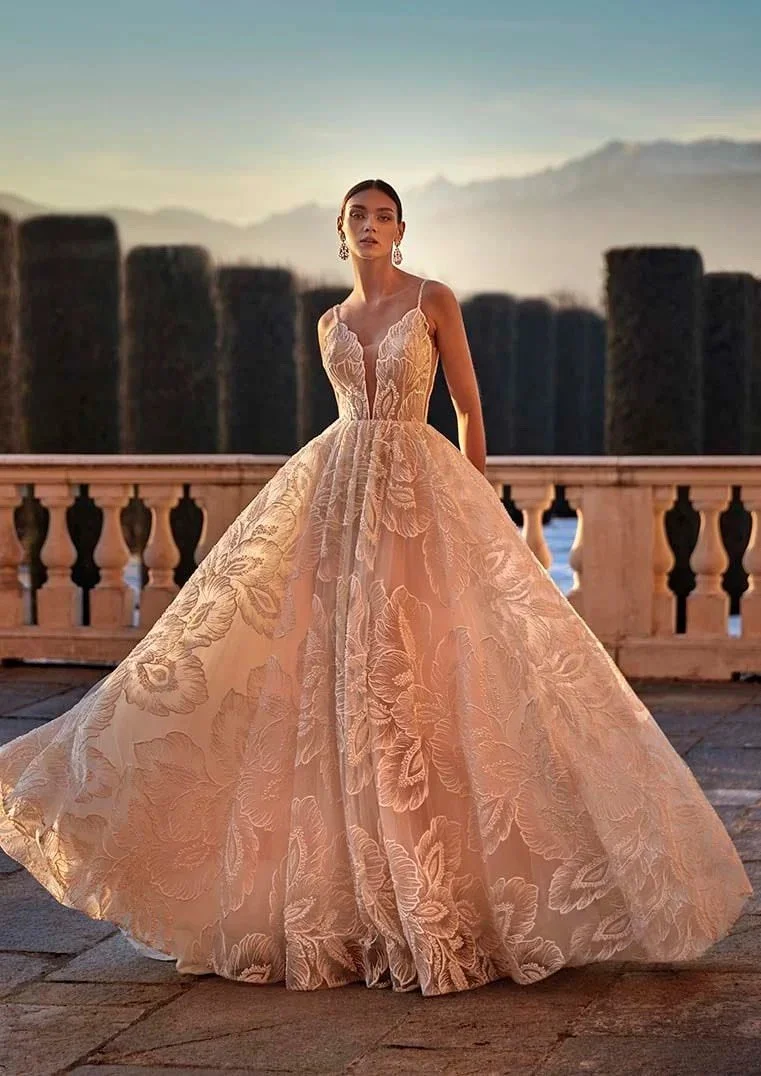A woman in a long, elegant floral lace gown standing outdoors on a balcony during sunset, with mountains in the background.