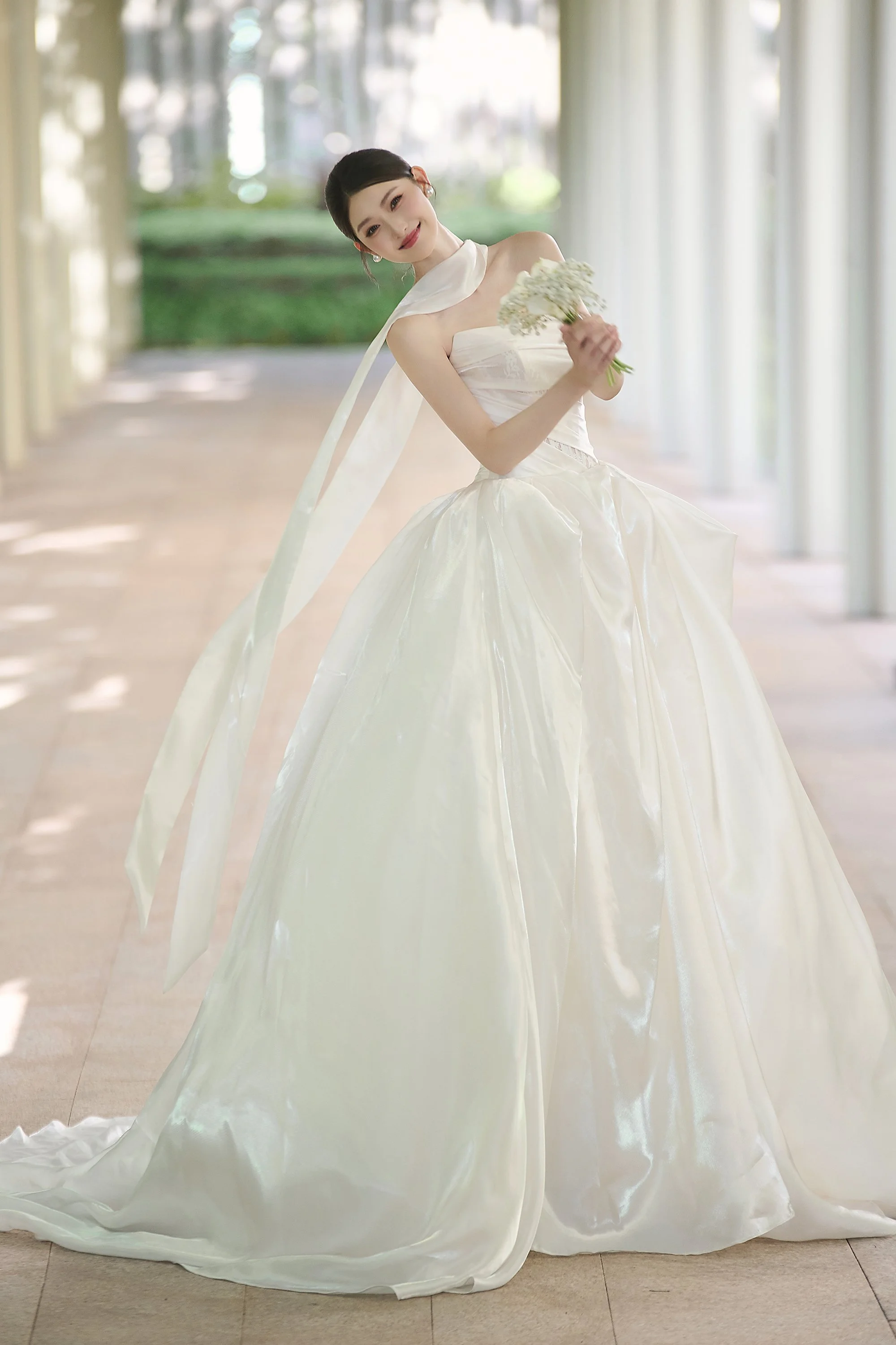 A woman in a white wedding gown holding a bouquet of flowers in a bright, airy corridor with large windows and natural light.