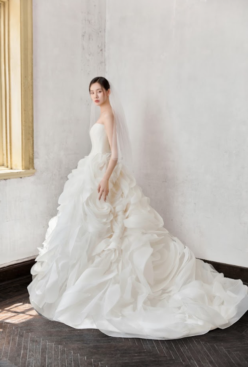 A bride in a strapless white wedding gown with ruffled layers standing indoors near a window with a draped veil.