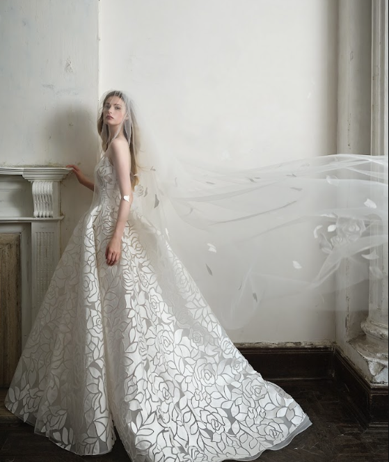 A woman in a white wedding dress with floral patterns, standing indoors near a wall, with a long veil flowing behind her.