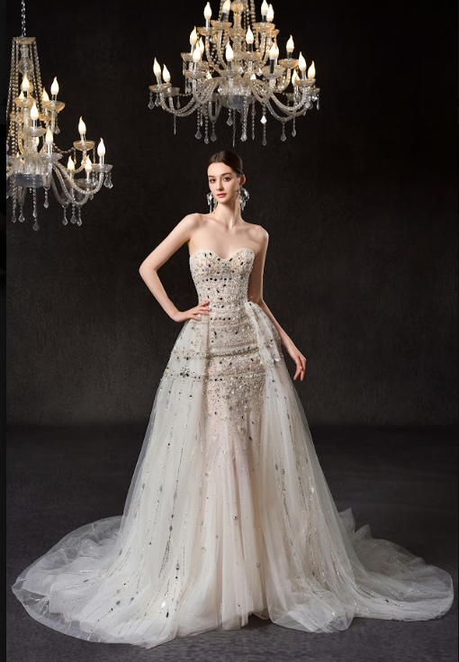 A woman in an elegant wedding gown standing under a chandelier with dark background.