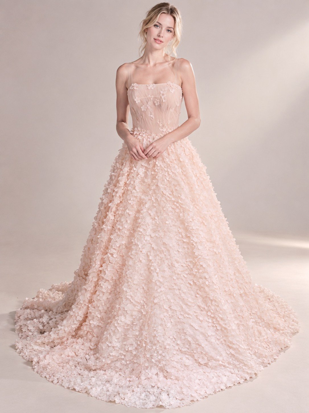 A woman wearing a light pink, floral textured evening gown with spaghetti straps, standing in a plain, light-colored studio background.