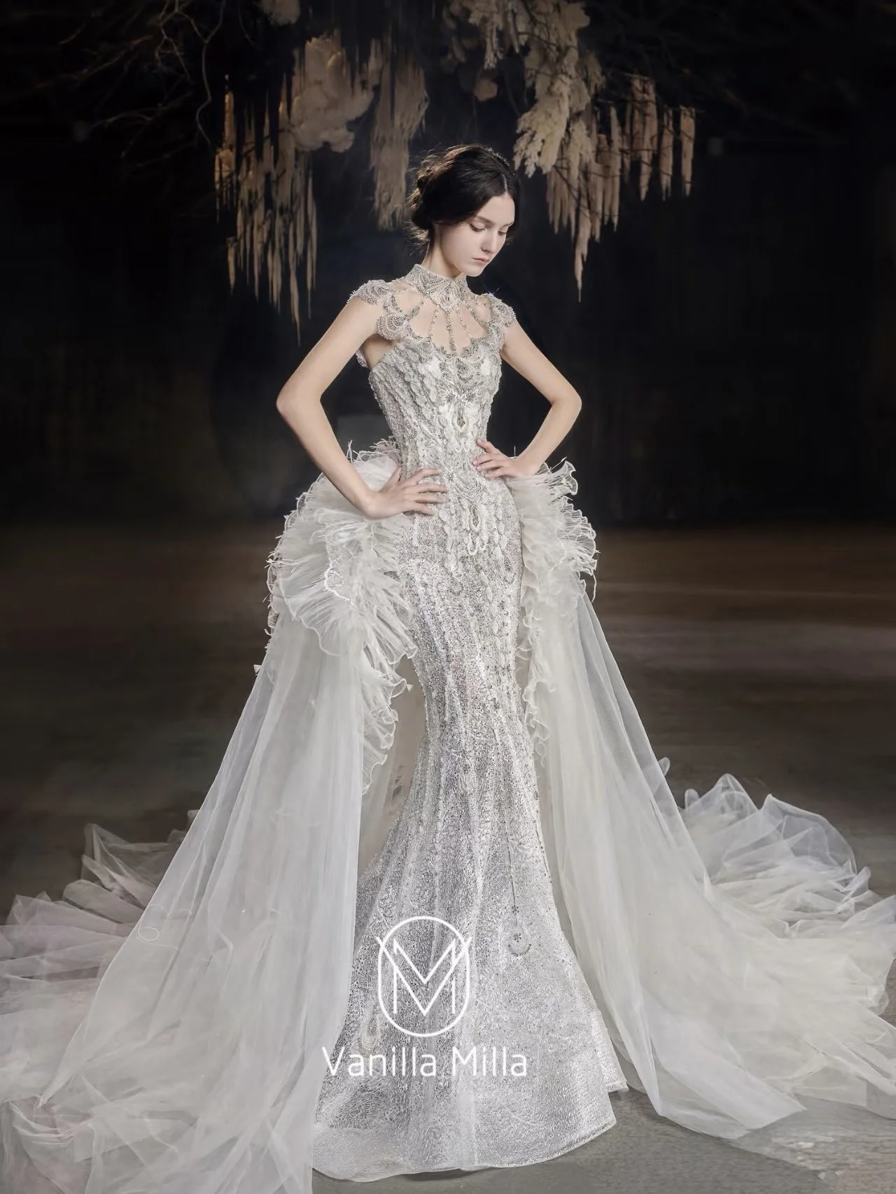 A woman in an elaborate, silver and white wedding gown with intricate lace and beadwork, standing with her hands on her hips, in a dark setting with hanging dried foliage in the background.