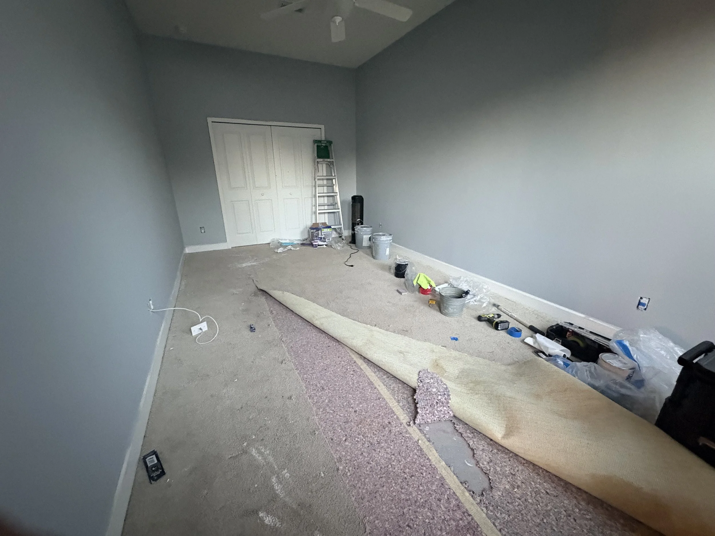 Room under renovation with walls painted gray, a ceiling fan, and various painting tools and supplies scattered on the carpet and floor.